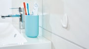 Een witte tesa Powerstrips-haak (Small) aan een badkamermuur, zijaanzicht met wastafel en tandenborstelhouder op de achtergrond.