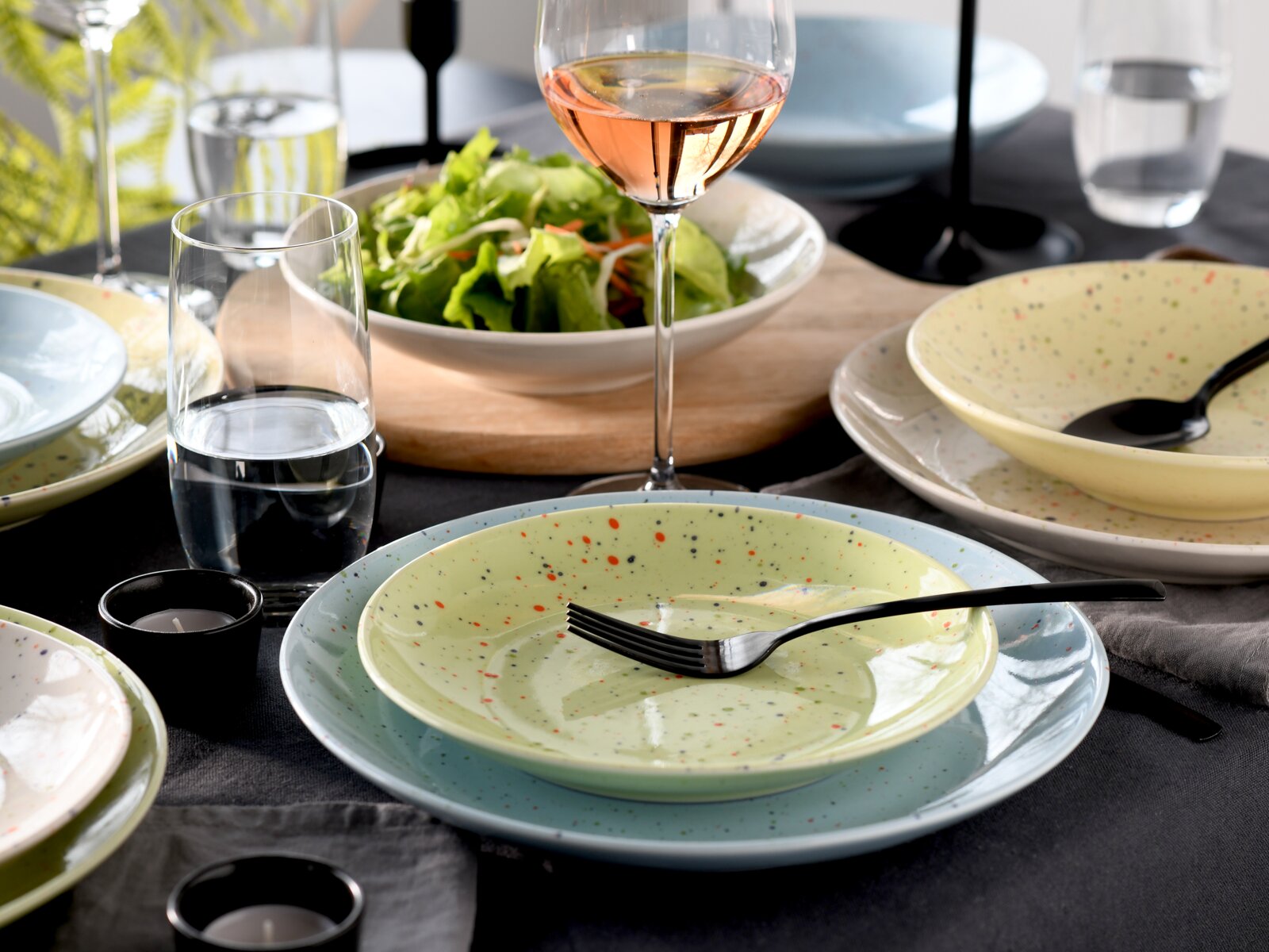 Modern gedeckter Esstisch mit gesprenkeltem Keramik-Geschirr in Pastellfarben (Teller und Schalen), schwarzem Besteck, Salatschüssel und Roséwein- sowie Wassergläsern; Perspektive: leicht schräg von oben auf das Tischgedeck.