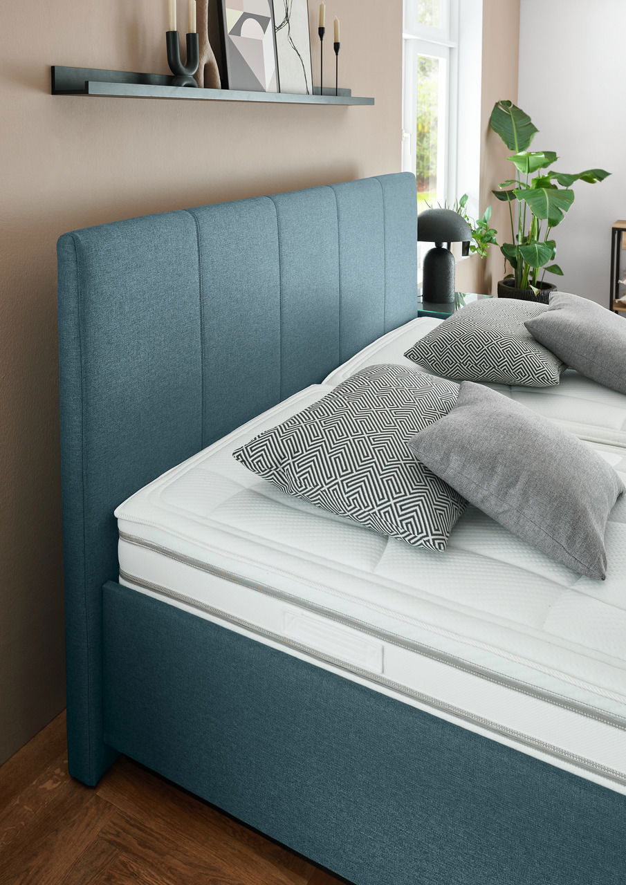 Seitliche Perspektive eines modernen Polsterbetts in Blau mit gestepptem Kopfteil, dekoriert mit gemusterten Kissen und umgeben von stilvollen Accessoires.