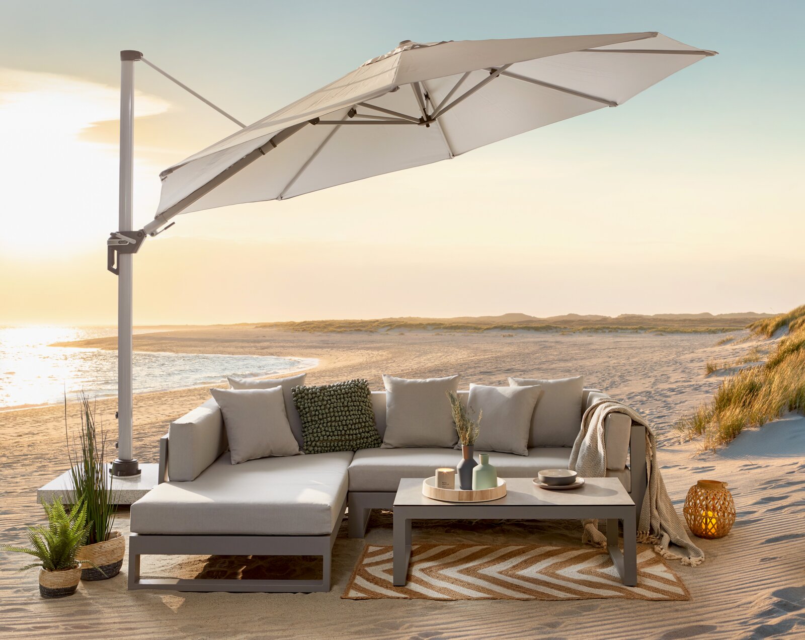 Heller Ampelschirm mit seitlichem Mast über einer modernen Outdoor-Lounge am Strand bei Sonnenuntergang, Perspektive: leicht seitliche Ansicht von vorn.
