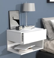 Weißer, wandmontierter Nachttisch mit geöffneter Schublade und Ablagefach, darauf eine silberne Tischlampe, Bücher, ein Wecker und ein gerahmtes Bild; Perspektive: frontale Ansicht leicht von links.