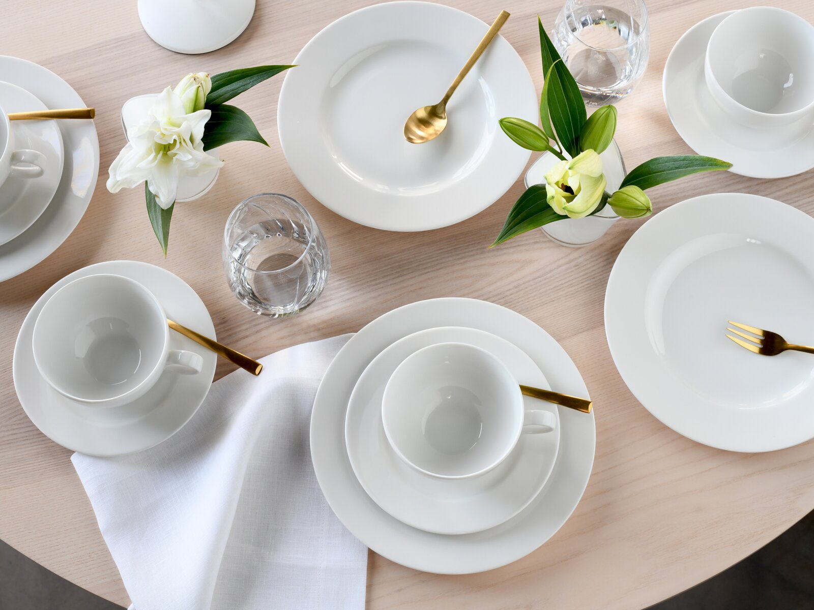 Draufsicht auf ein elegantes, weißes Porzellan-Geschirrset mit Tellern, Tassen und Schalen auf hellem Holztisch, ergänzt durch goldfarbenes Besteck, Wassergläser, weiße Serviette und Blumenarrangement.
