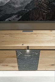 Frontale Ansicht einer Wohnwand mit Holz- und Steinelementen, darunter eine Schublade und eine Tür mit Beleuchtung.