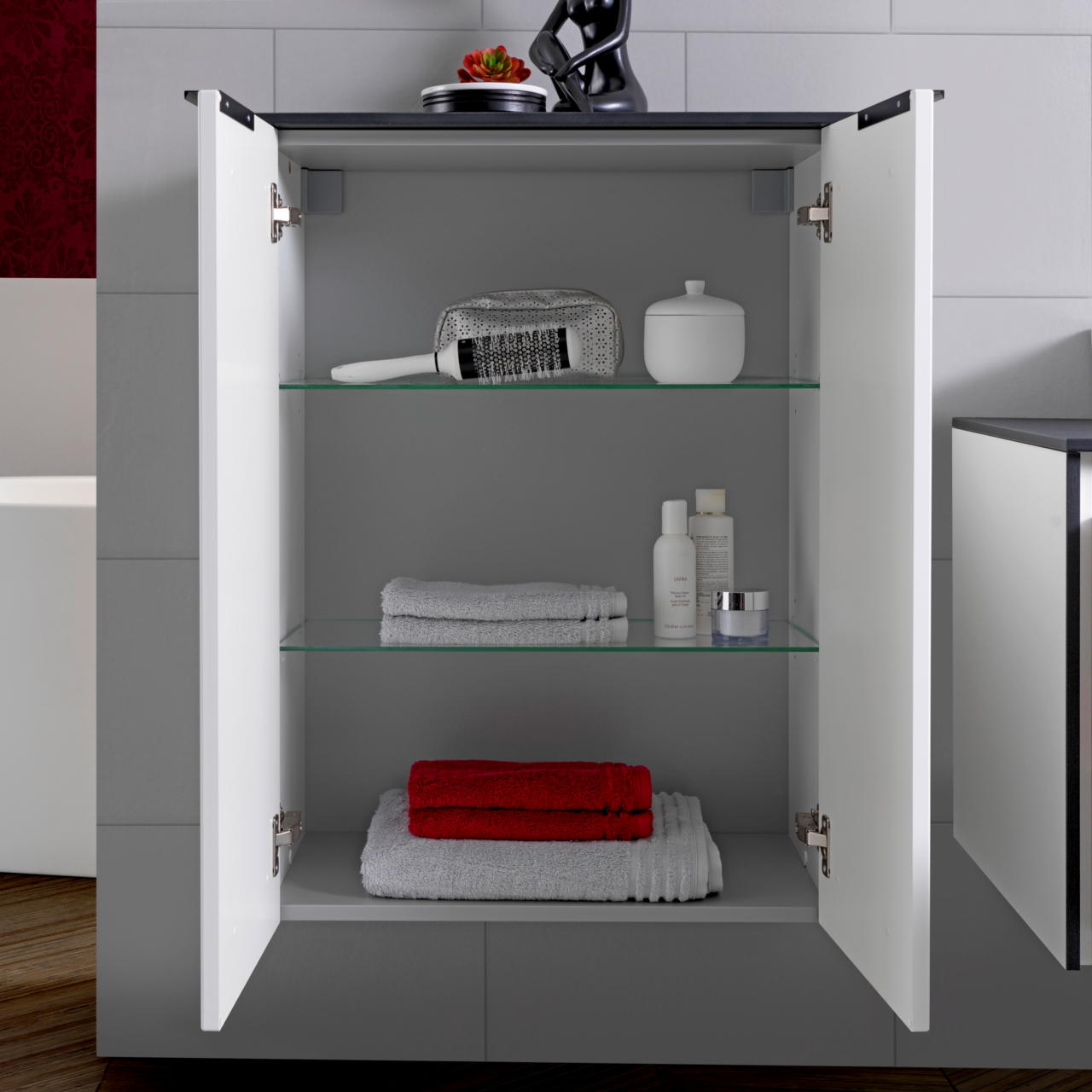 Offenes Highboard mit zwei Glaseinlegeböden, gefüllt mit Handtüchern und Pflegeprodukten, frontal betrachtet.