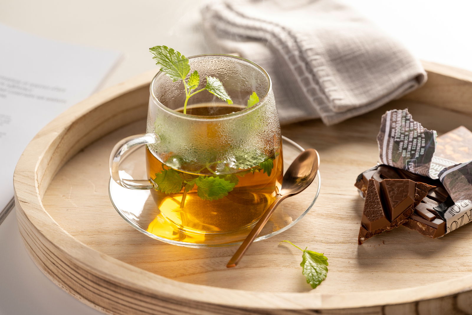 Glasuntertasse mit Teetasse, gefüllt mit Kräutertee, von oben fotografiert, auf einem Holztablett mit Schokolade und Löffel.