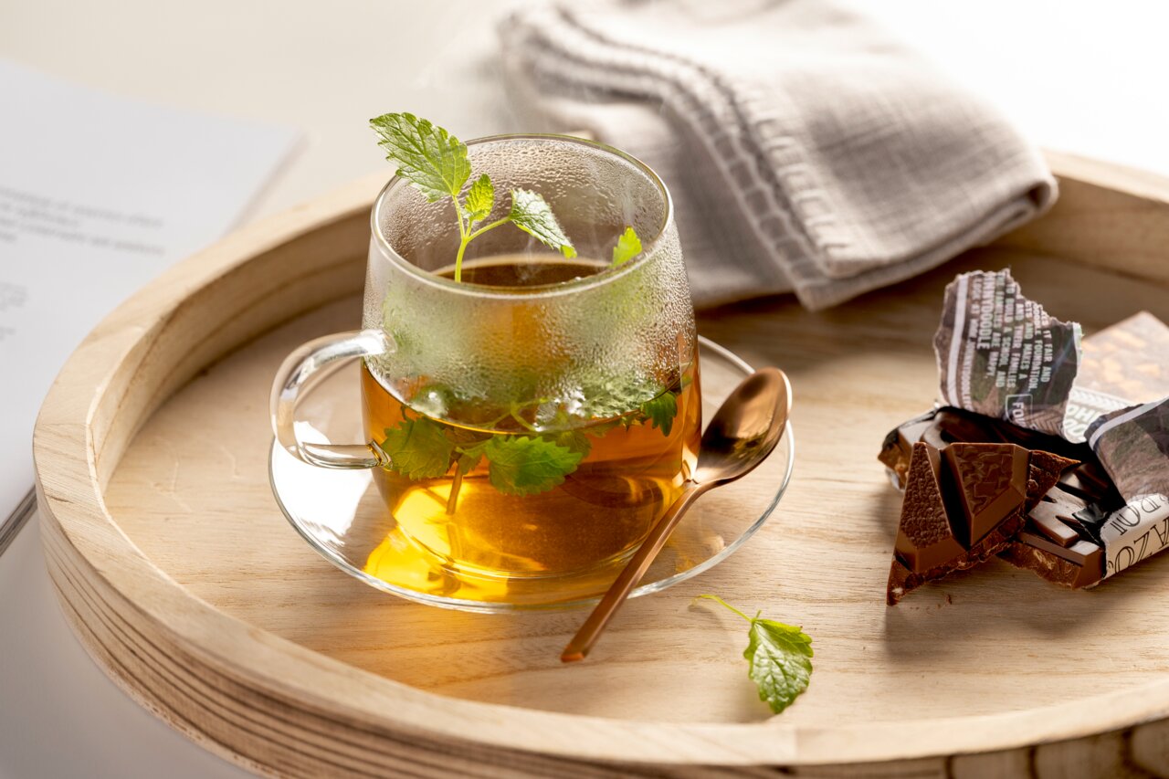 Glasuntertasse mit Teetasse, gefüllt mit Kräutertee, von oben fotografiert, auf einem Holztablett mit Schokolade und Löffel.