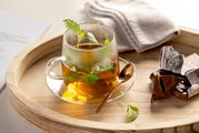 Glasuntertasse mit Teetasse, gefüllt mit Kräutertee, von oben fotografiert, auf einem Holztablett mit Schokolade und Löffel.