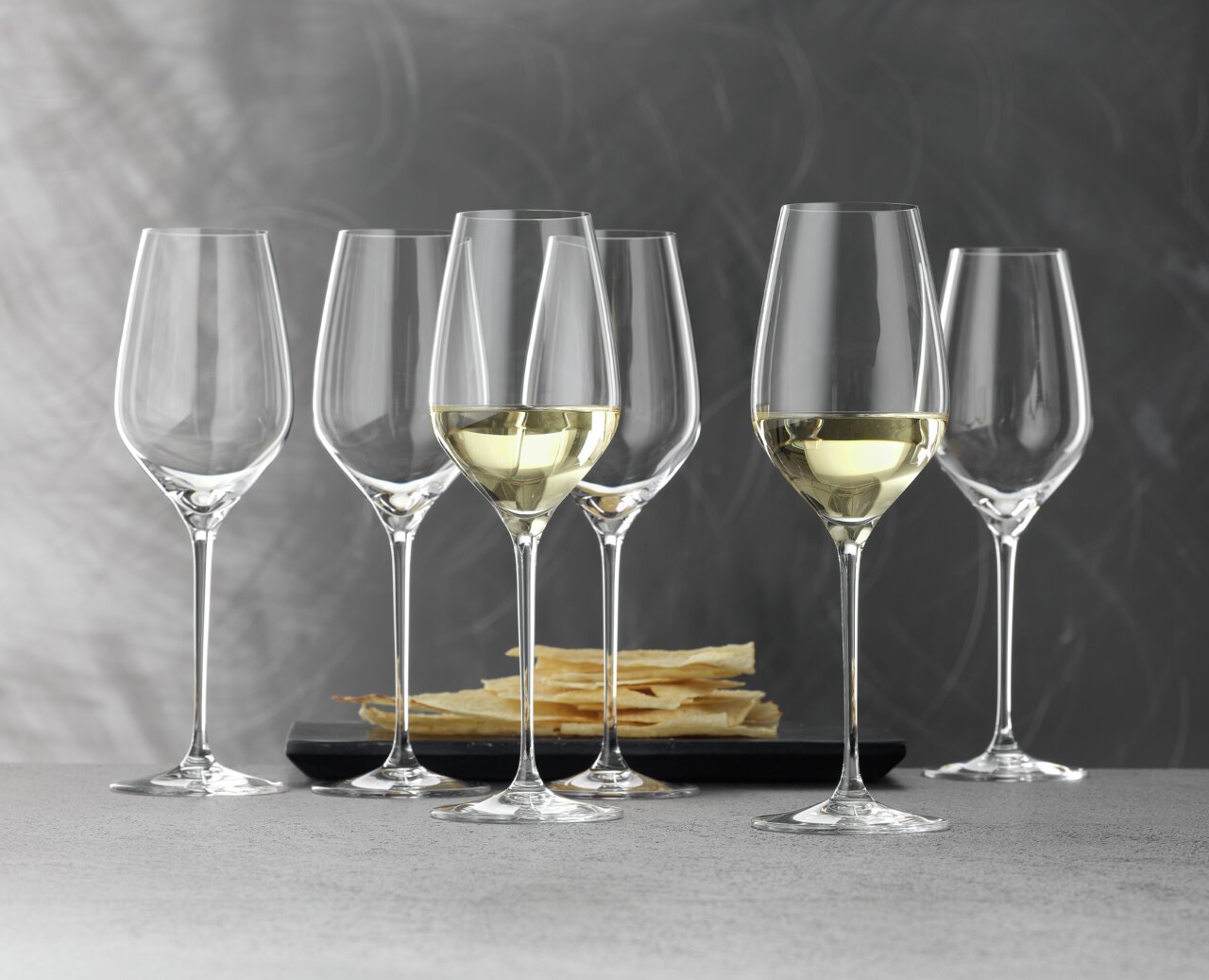 Sechs elegante Weißweinkelche der Serie Topline, teilweise gefüllt, auf einem Tisch mit Knabbergebäck im Hintergrund, seitliche Perspektive.