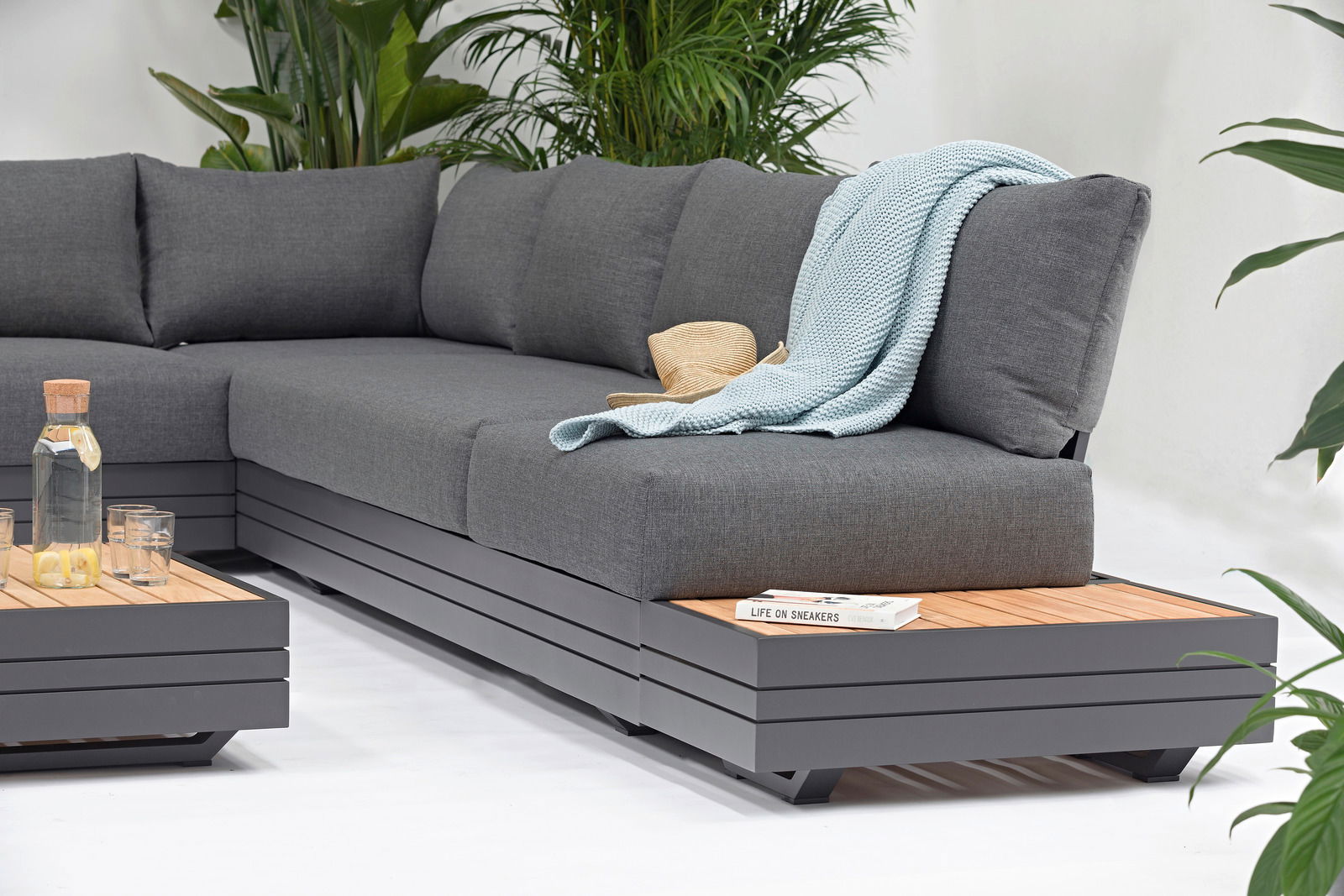 Graues Lounge-Sofa mit Kissen, Decke und Hut, umgeben von Pflanzen, seitliche Perspektive