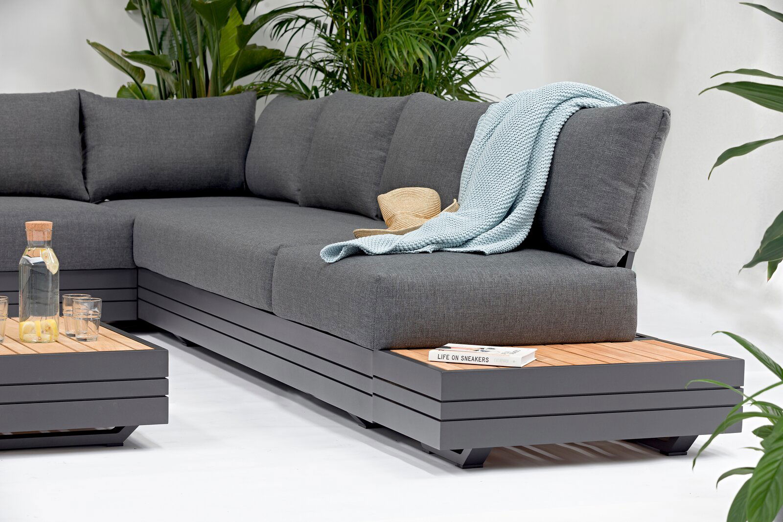 Graues Lounge-Sofa mit Kissen, Decke und Hut, umgeben von Pflanzen, seitliche Perspektive
