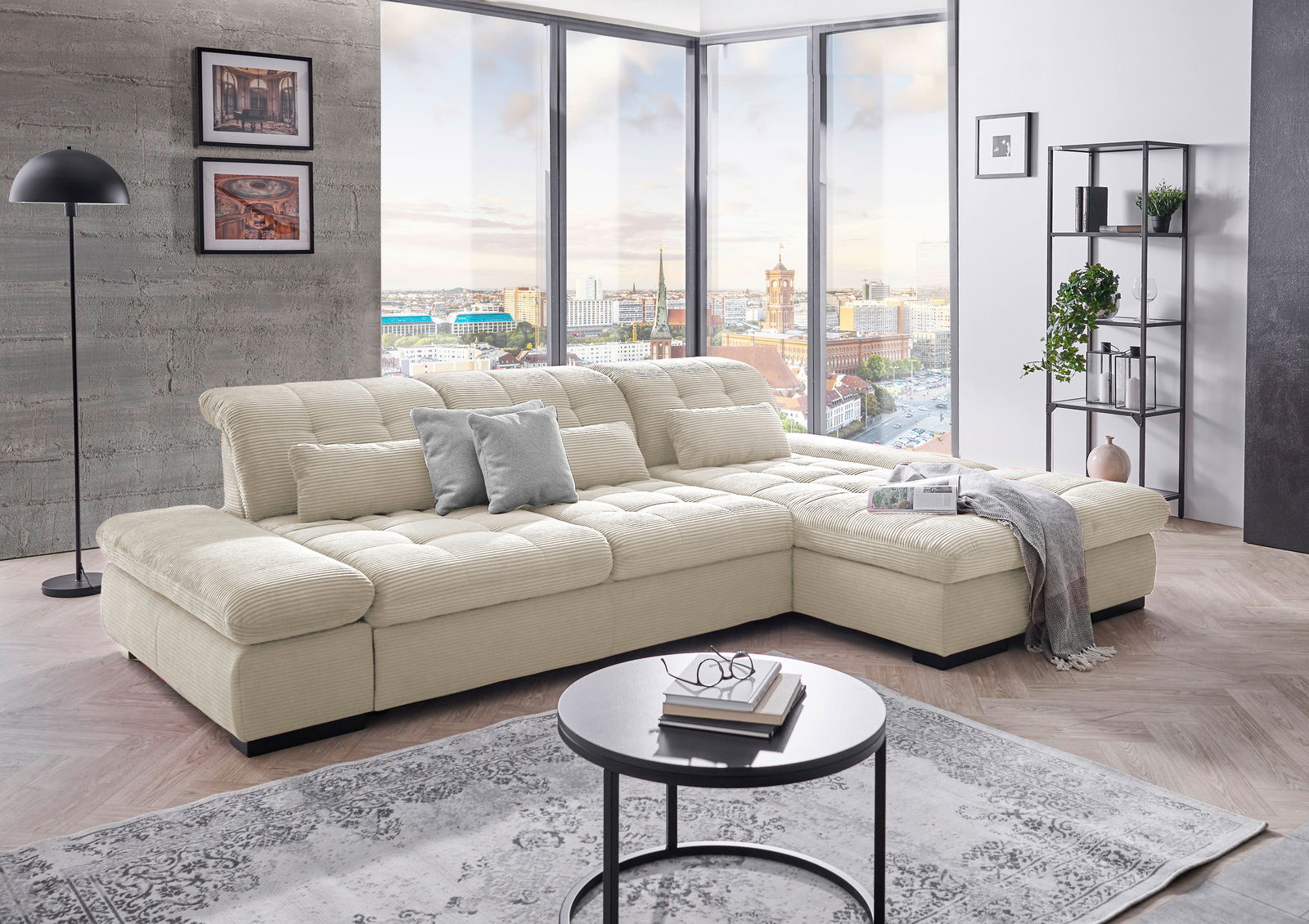 Großes, beiges Ecksofa in einem modernen Wohnzimmer mit Panoramafenstern, die einen Stadtblick bieten. Perspektive von vorne links.