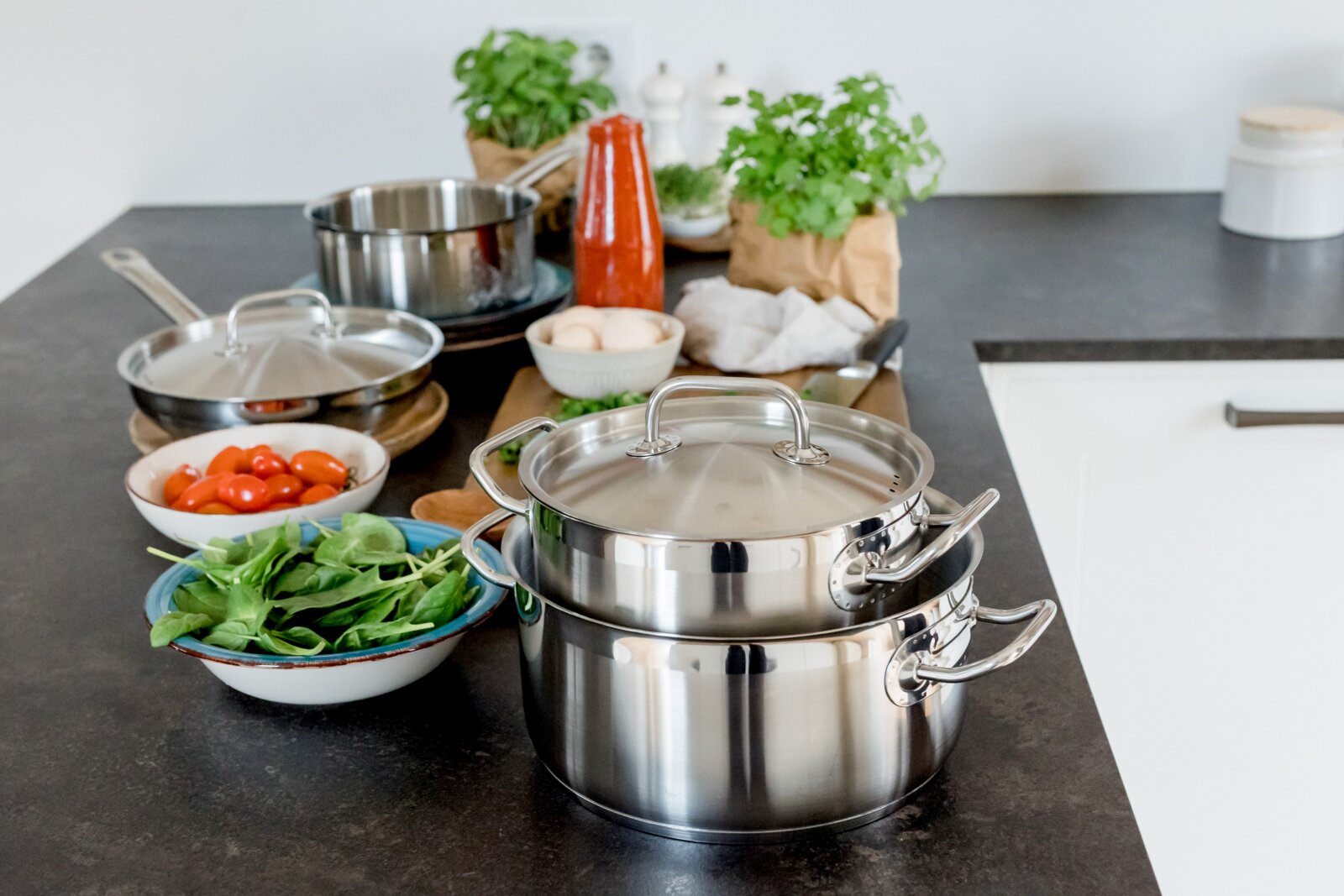 Edelstahl-Kochtopf mit Deckel auf Küchenarbeitsplatte, im Vordergrund mit leicht seitlicher Perspektive; im Hintergrund Zutaten wie Spinat, Tomaten, Eier, Kräuter und eine Flasche Tomatensauce