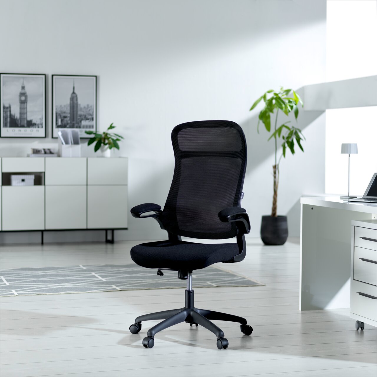 Moderner schwarzer Drehstuhl mit Netzrückenlehne und Armlehnen, seitliche Perspektive in einem hellen Büro mit Pflanzen und Dekoration.