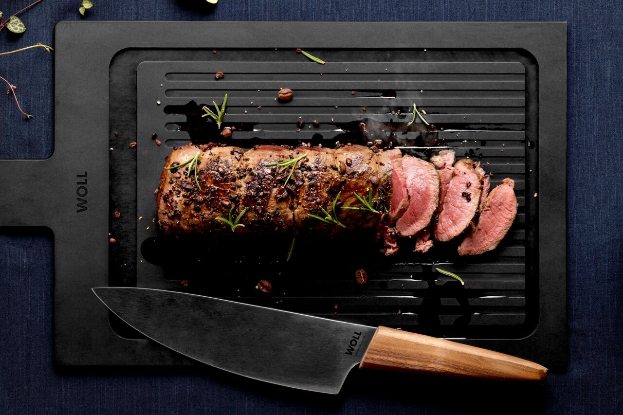 Woll Schneidebrett mit Rillen CULT & SERVE Vogelperspektive auf ein Schneidebrett mit Rillen, darauf ein gebratenes und in Scheiben geschnittenes Stück Fleisch, garniert mit Rosmarin, daneben ein Messer mit Holzgriff.