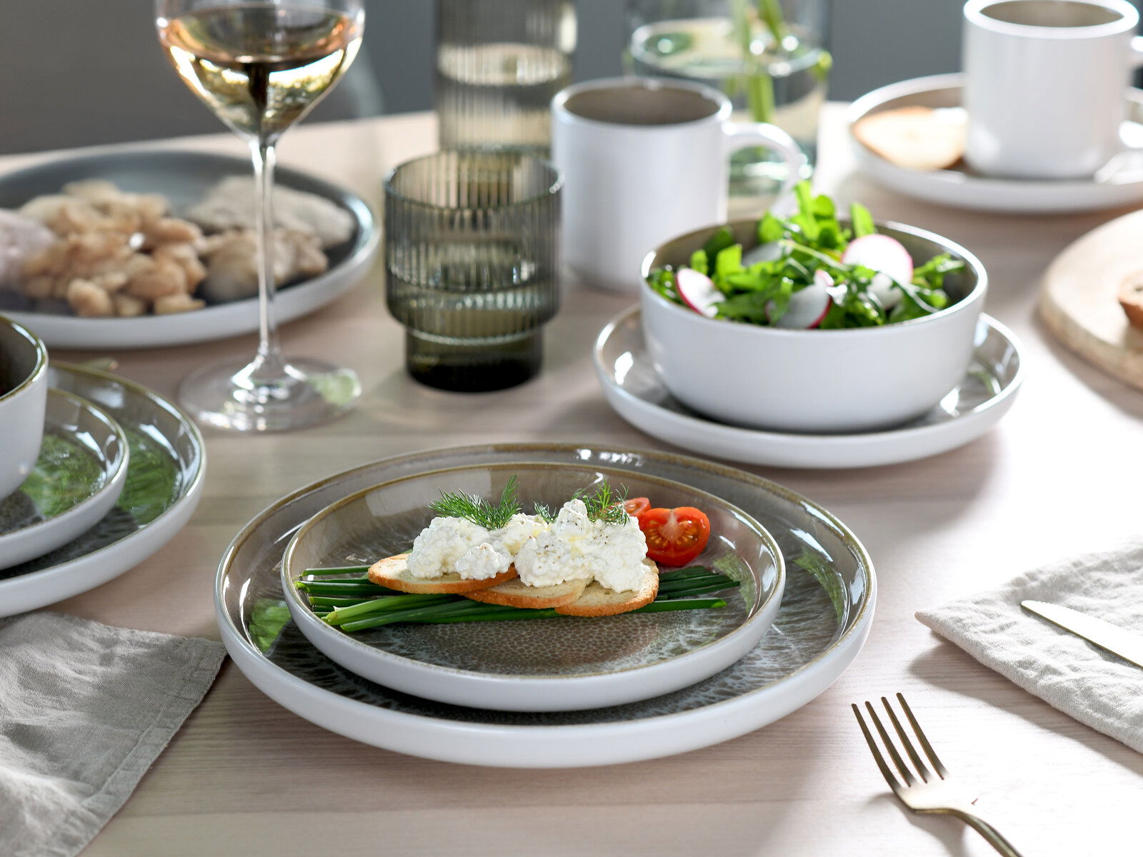 Ein stilvoll gedeckter Tisch mit einem 16-teiligen Kombiservice in Sandfarbe, bestehend aus Tellern, Schalen und Tassen. Im Vordergrund ein Teller mit Brot, Frischkäse und Gemüse. Perspektive von oben.