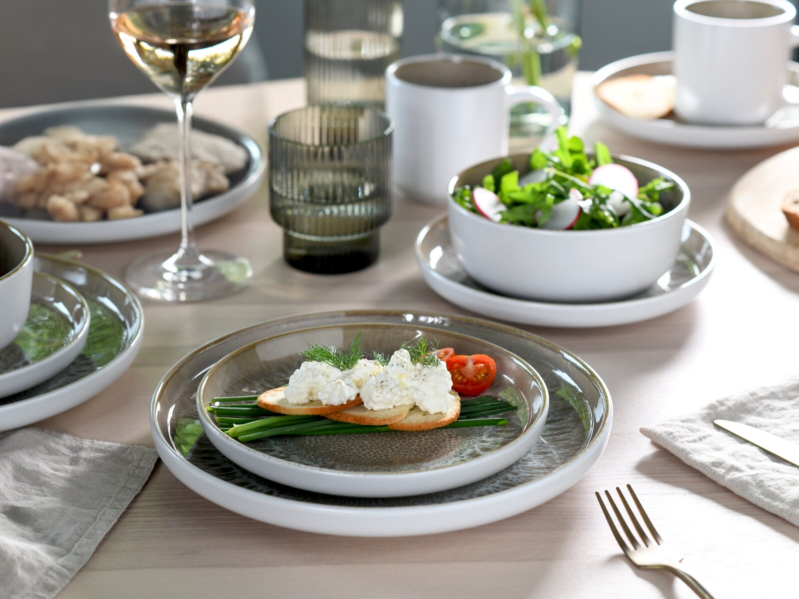 Ein stilvoll gedeckter Tisch mit einem 16-teiligen Kombiservice in Sandfarbe, bestehend aus Tellern, Schalen und Tassen. Im Vordergrund ein Teller mit Brot, Frischkäse und Gemüse. Perspektive von oben.