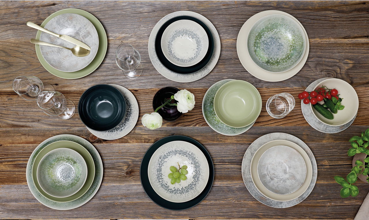 Vogelperspektive auf ein 18-teiliges Geschirrset in Graphit- und Grüntönen, arrangiert auf einem Holztisch mit Gläsern, Besteck und Dekoration.
