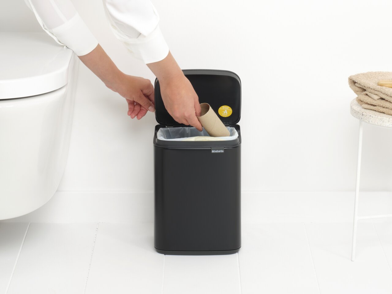 Brabantia Abfallbehälter 4L BO Schwarzer Abfallbehälter Bo 4L in einem Badezimmer, von der Seite gesehen, mit einer Hand, die einen Papprollenkern hineinwirft.