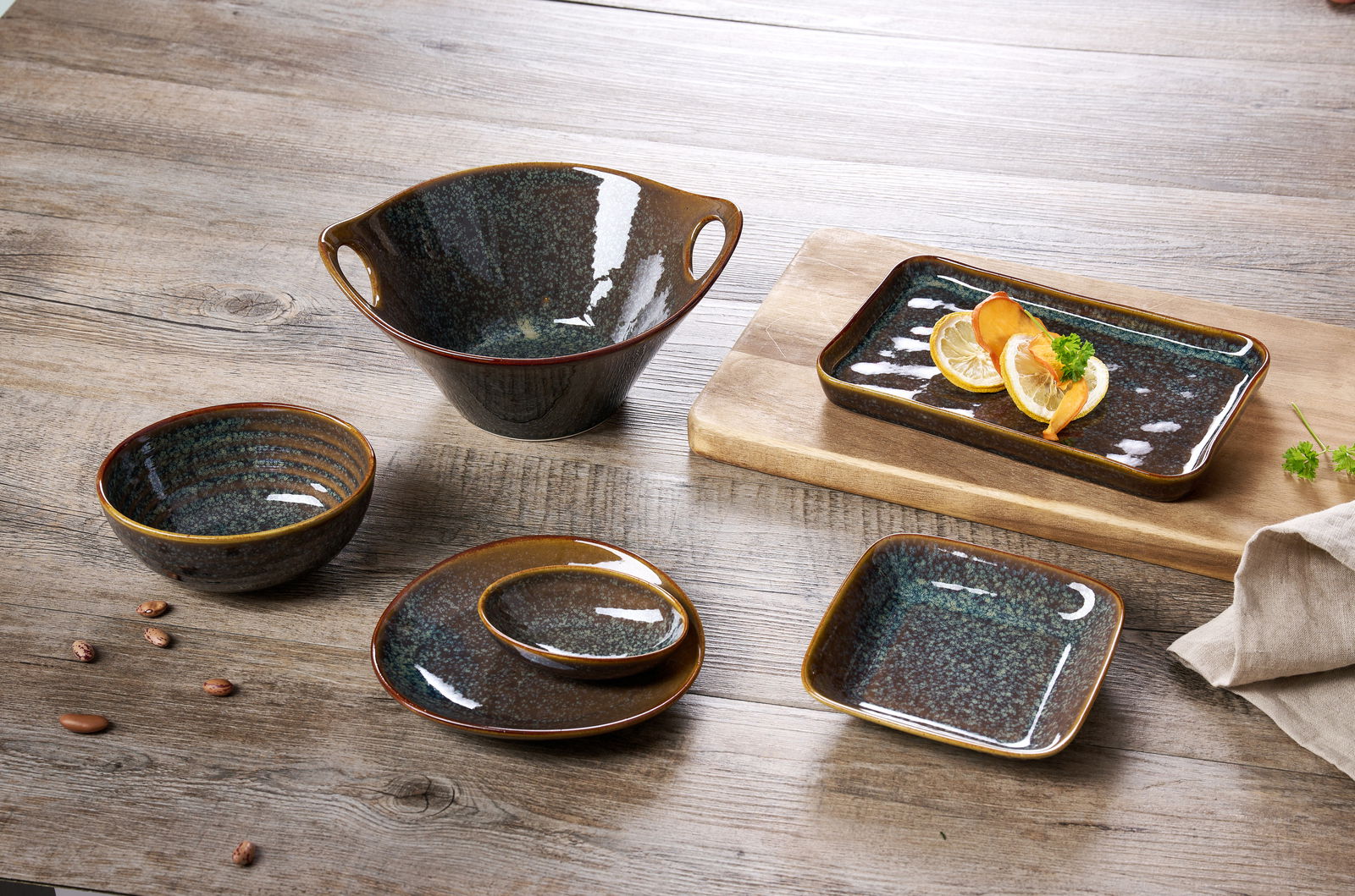 Ritzenhoff & Breker Beilagenteller TROMS Keramikgeschirr-Set auf einem Holztisch, bestehend aus einer großen Schale mit Henkeln, einer rechteckigen Auflaufform mit Zitronenscheiben, einer runden Schale, einem ovalen Teller mit kleiner Schale und einem quadratischen Teller. Perspektive von oben.