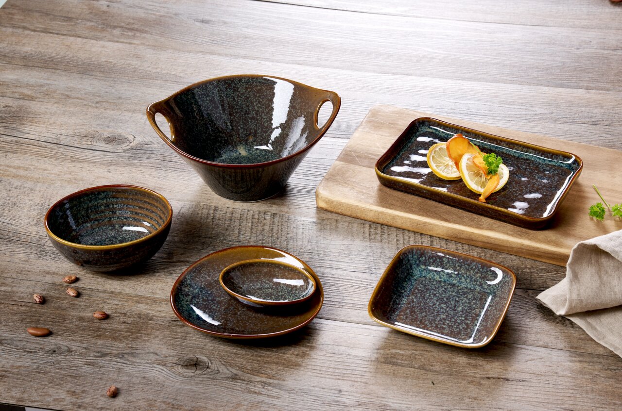 Ritzenhoff & Breker Beilagenteller TROMS Keramikgeschirr-Set auf einem Holztisch, bestehend aus einer großen Schale mit Henkeln, einer rechteckigen Auflaufform mit Zitronenscheiben, einer runden Schale, einem ovalen Teller mit kleiner Schale und einem quadratischen Teller. Perspektive von oben.