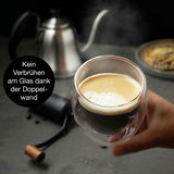 Nahaufnahme eines doppelwandigen Glases mit heißem Kaffee, gehalten in einer Hand. Im Hintergrund eine silberne Kanne. Kein Verbrühen am Glas dank der Doppelwand.