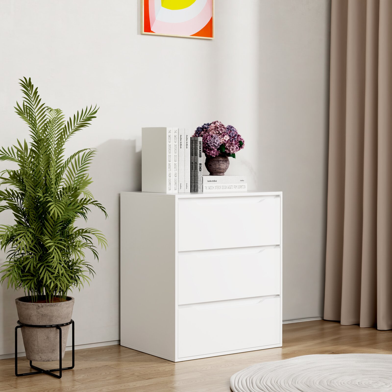Weiße, moderne Kommode mit drei Schubladen in einer hellen Zimmerecke, oben mit Büchern und Blumenvase dekoriert Frontansicht in leichter Schrägperspektive von links.