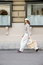 Seitenansicht: Frau geht auf dem Gehweg und trägt einen großen beigefarbenen Korb mit Muster und Ledergriffen vor einer Hausfassade mit Fenstern.