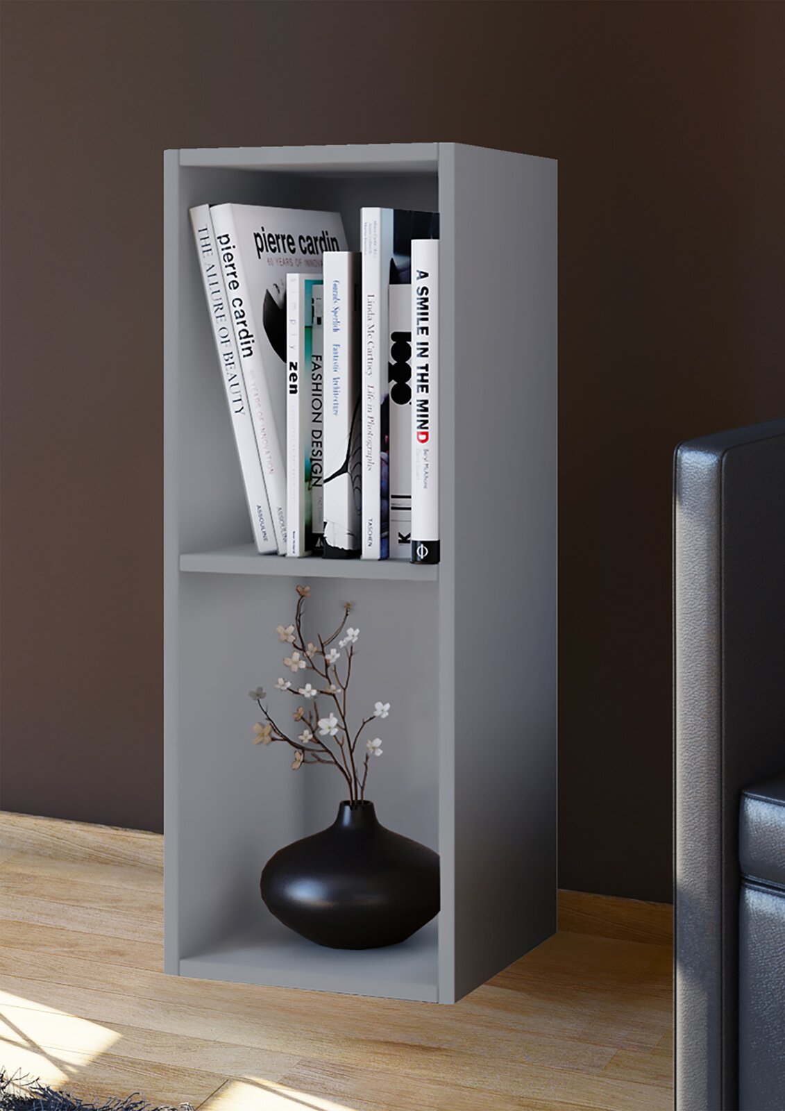 Graues, schmales Standregal mit zwei offenen Fächern, oben mit Büchern und unten mit schwarzer Vase samt Zweig, in leicht schräger Frontansicht im Wohnraum neben einem Sofa