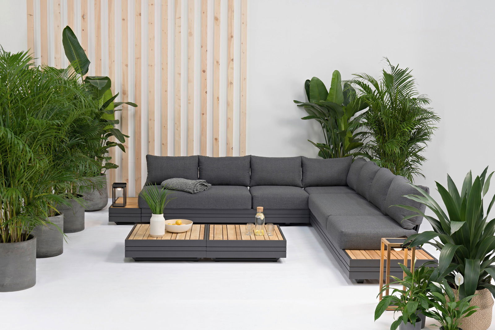 Zebra Loungestoel CUBO Moderne Gartenmöbel-Lounge mit grauem Ecksofa und Holztischen, umgeben von grünen Pflanzen, aus der Vorderansicht.