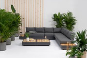 Moderne Gartenmöbel-Lounge mit grauem Ecksofa und Holztischen, umgeben von grünen Pflanzen, aus der Vorderansicht.