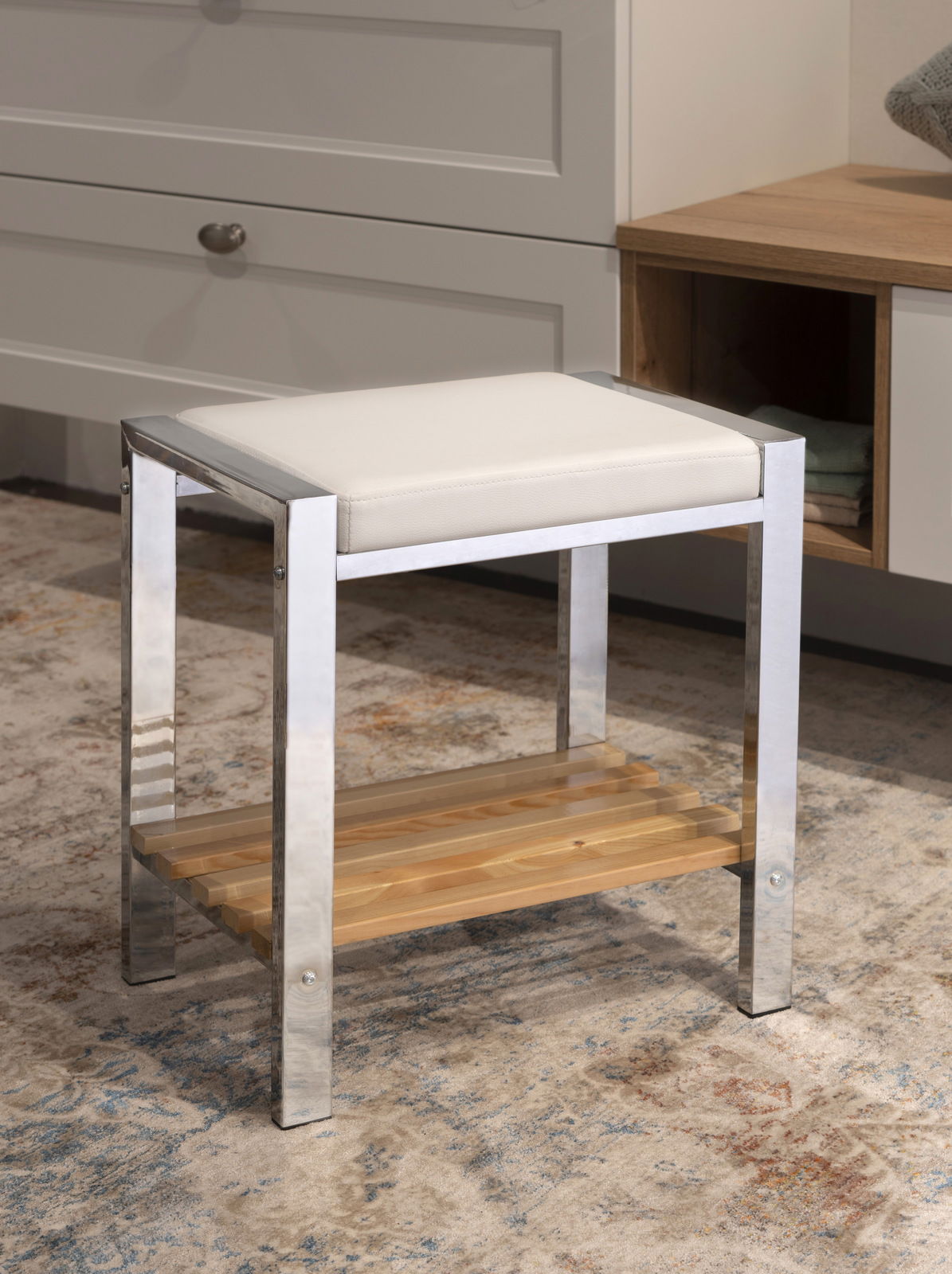 Moderner Hocker mit weißem Sitzpolster und Metallrahmen, darunter eine Holzablage. Perspektive: seitliche Ansicht.