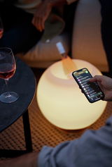 Eine leuchtende, kugelförmige Lampe in gebürstetem Silber, die als stilvolle Tischdekoration dient. Perspektive von oben, mit einem Smartphone und einem Weinglas im Vordergrund.