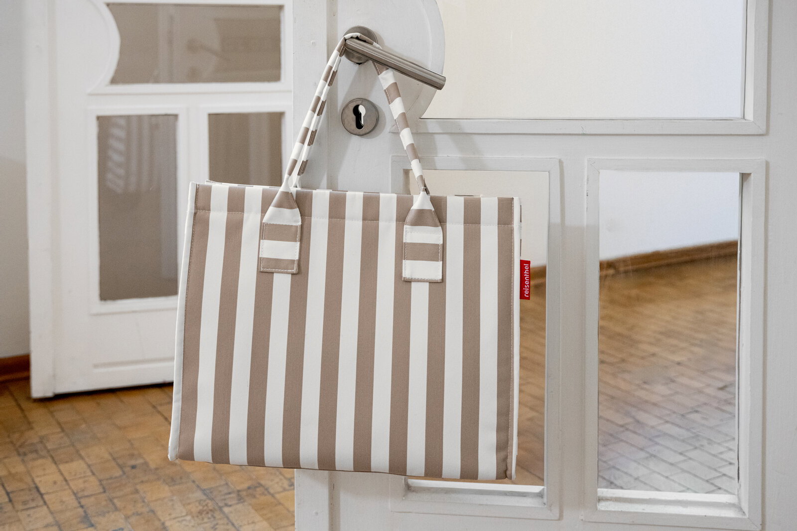 Beige-weiß gestreifte Tragetasche mit zwei Henkeln hängt an einer weißen Tür mit Metallklinke, frontale Ansicht in leicht schräger Perspektive.