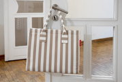 Een beige-wit gestreepte draagtas met twee hengsels hangt aan een witte deur met een metalen klink; vooraanzicht vanuit een licht schuin perspectief.