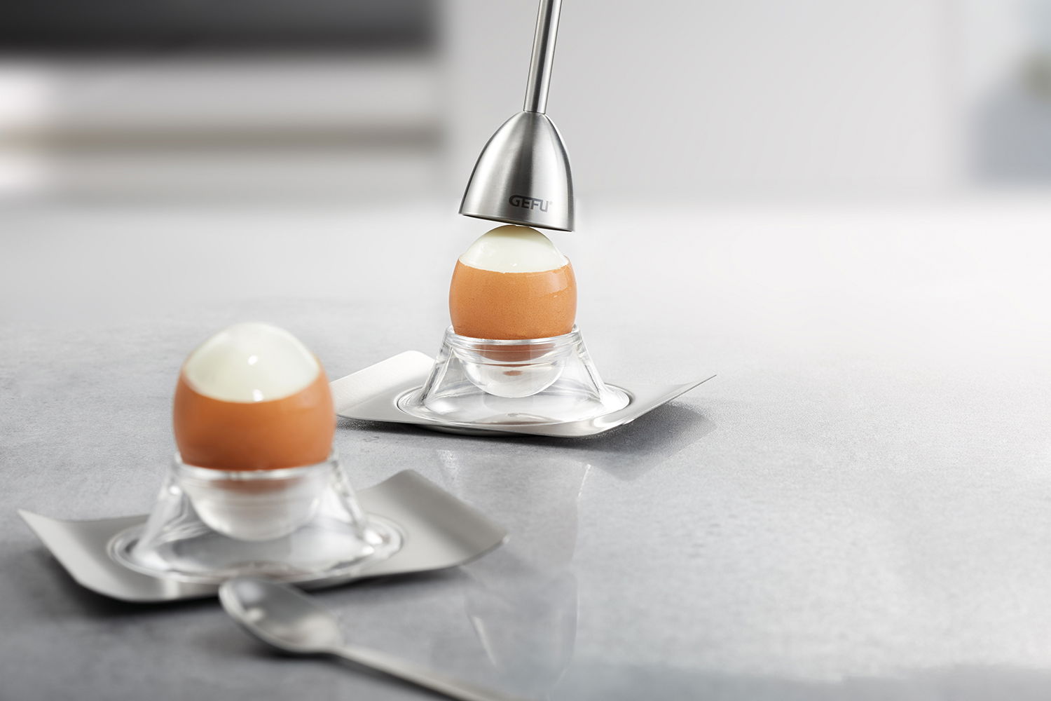 Eierköpfer mit Salzstreuer in moderner Küche, seitliche Perspektive auf zwei gekochte Eier in Eierbechern, eines wird mit dem Eierköpfer geöffnet.