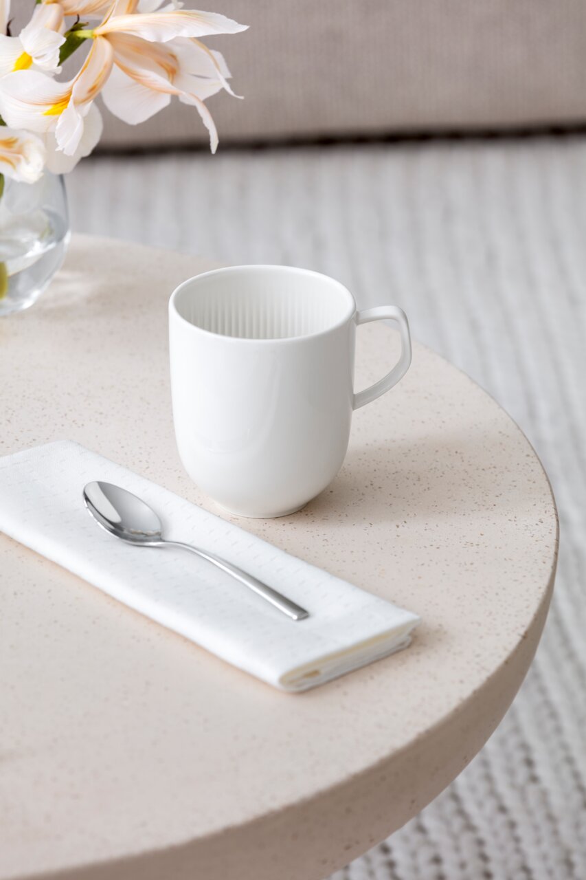 Villeroy & Boch Kaffeebecher AFINA Weißer Becher mit Henkel auf einem runden Tisch, neben einer Serviette und einem Löffel, aus der Vogelperspektive fotografiert.