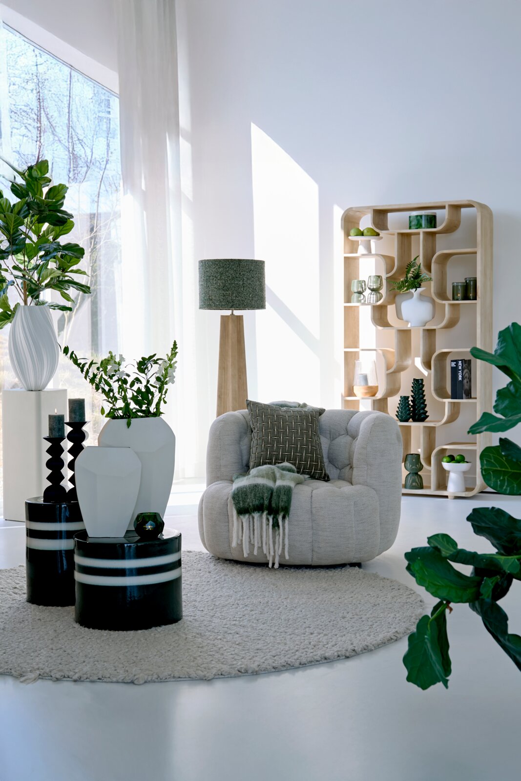 Light&Living Vrijstaande boekenkast LABARE Moderne houten standaard met gebogen planken, versierd met planten en vazen, in een lichte woonkamer met grote ramen, gezien vanuit de zijkant.