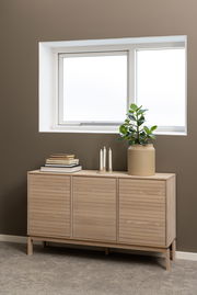 Modernes Sideboard aus hellem Holz mit drei Türen, dekoriert mit Büchern, Kerzen und einer Pflanze, seitliche Perspektive.