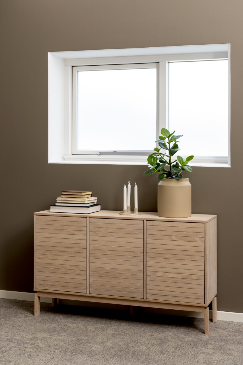 Modernes Sideboard aus hellem Holz mit drei Türen, dekoriert mit Büchern, Kerzen und einer Pflanze, seitliche Perspektive.