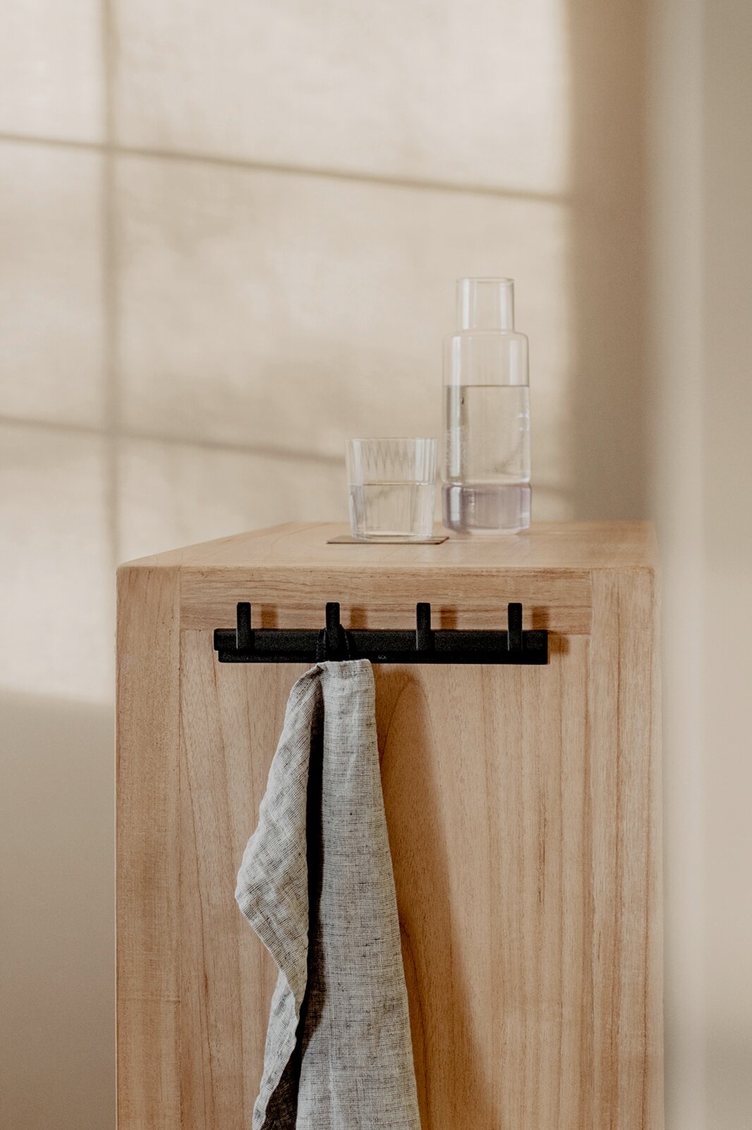 Frontansicht eines hellen Holz-Blocks mit schwarzer Hakenleiste und hängendem grauen Tuch; oben stehen ein Glas und eine klare Flasche, im Hintergrund ein sonnendurchflutetes Fenster.
