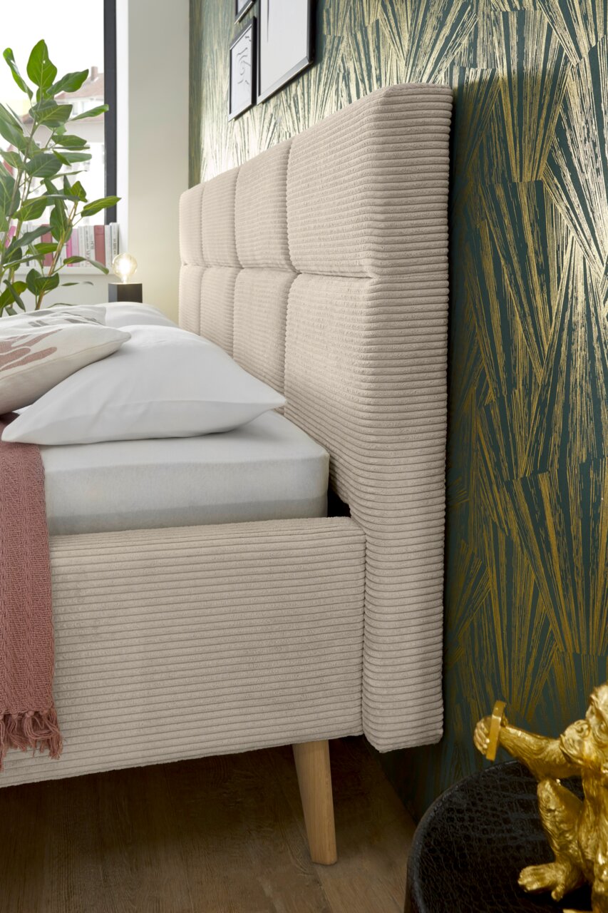 Seitliche Perspektive eines Polsterbetts mit beigem, geripptem Stoffbezug und Holzfüßen, vor einer grünen Tapete mit goldenen Akzenten.