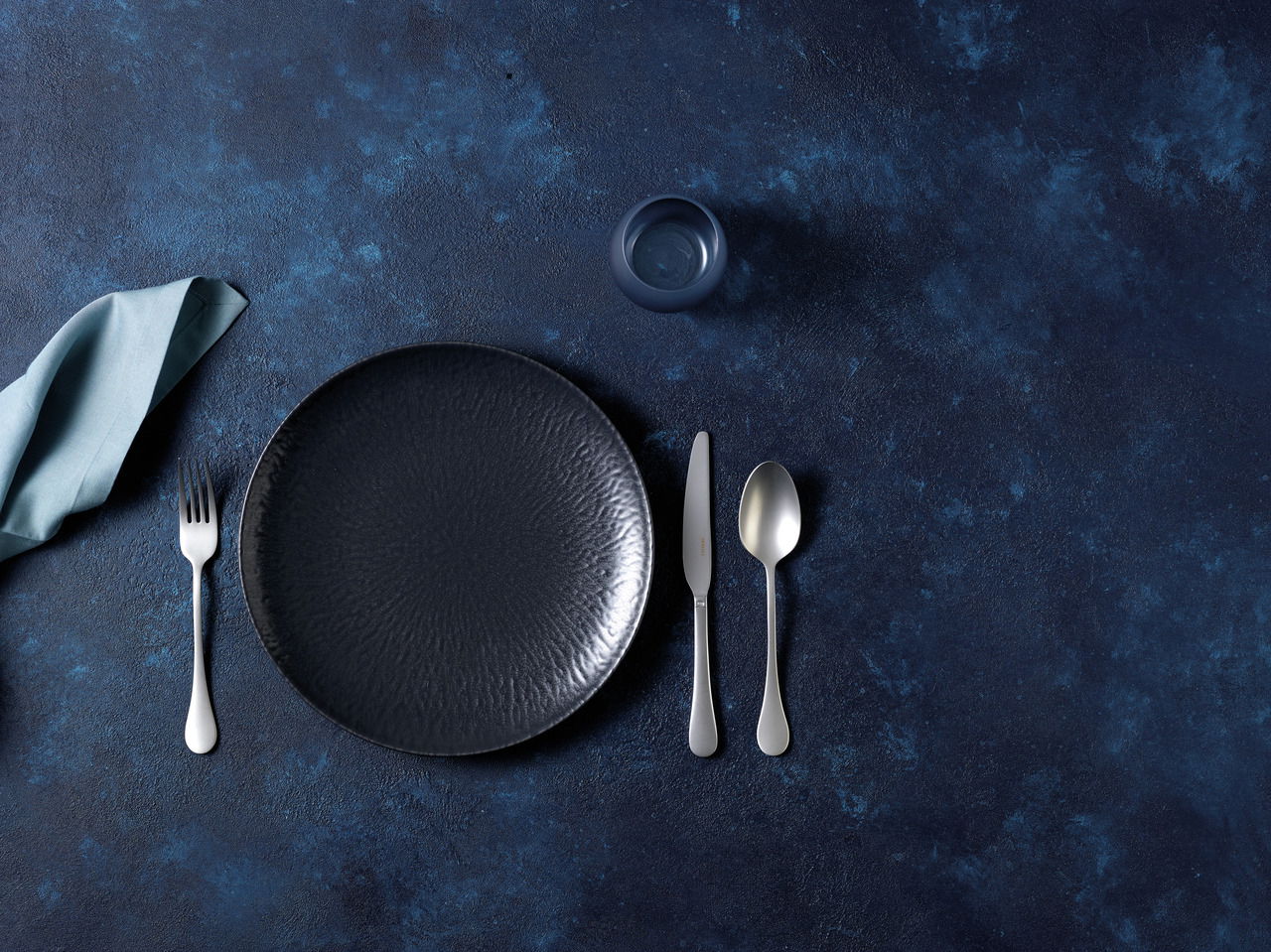 Arthur Krupp Tellerset 12-tlg. Onyx Draufsicht auf ein elegantes, schwarzes Tellerset mit Besteck und Serviette auf einem dunkelblauen Hintergrund.