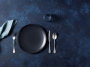 Draufsicht auf ein elegantes, schwarzes Tellerset mit Besteck und Serviette auf einem dunkelblauen Hintergrund.
