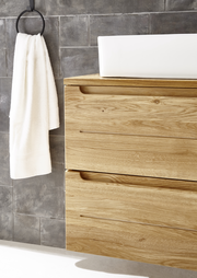 Moderner Waschtischunterschrank aus Holz mit zwei Schubladen, seitliche Perspektive, neben einem Handtuchhalter an einer grauen Wand.