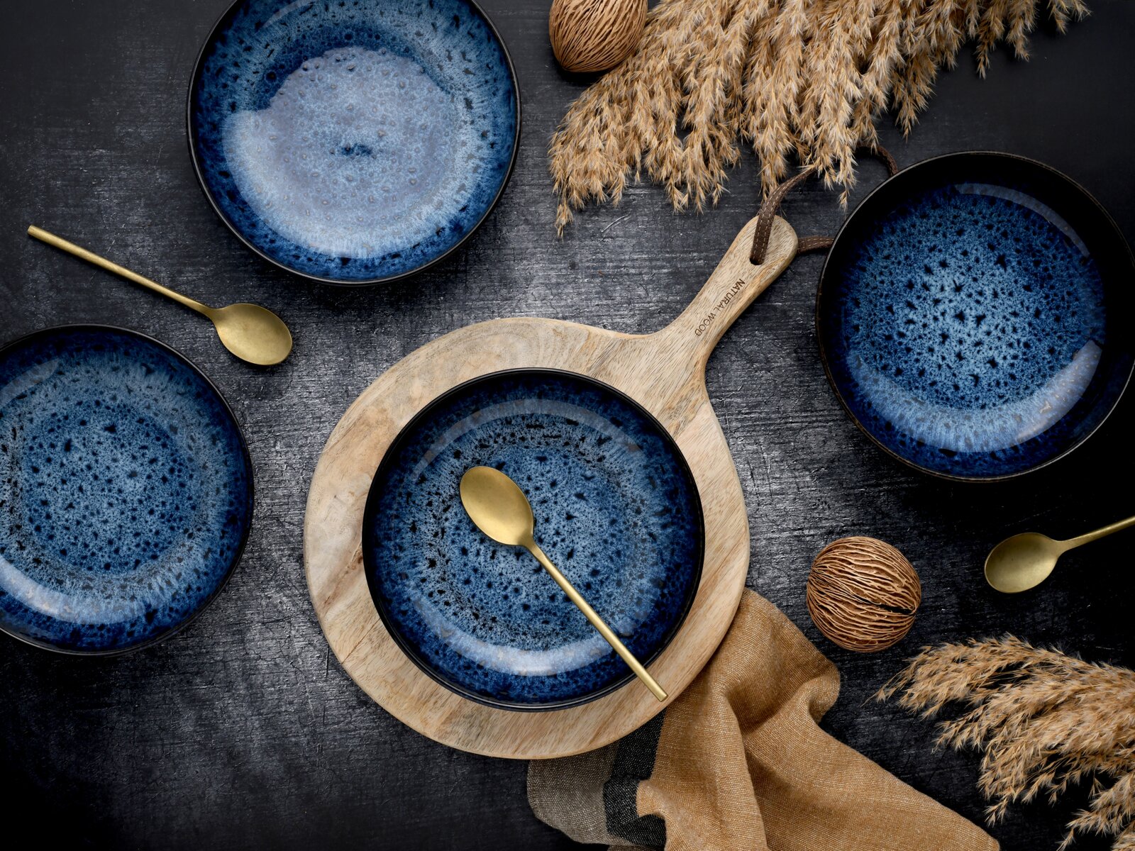 Dunkelblaue, gesprenkelte Keramikschalen mit goldfarbenen Löffeln auf rustikalem Holztisch, arrangiert um ein helles Holz-Servierbrett mit Griff – Draufsicht (Flatlay).