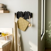 Moderne Wandgarderobe mit schwarzen Haken, an der ein beiger Stoffbeutel, ein gelber Topflappen und eine Spülbürste hängen. Perspektive von der Seite.