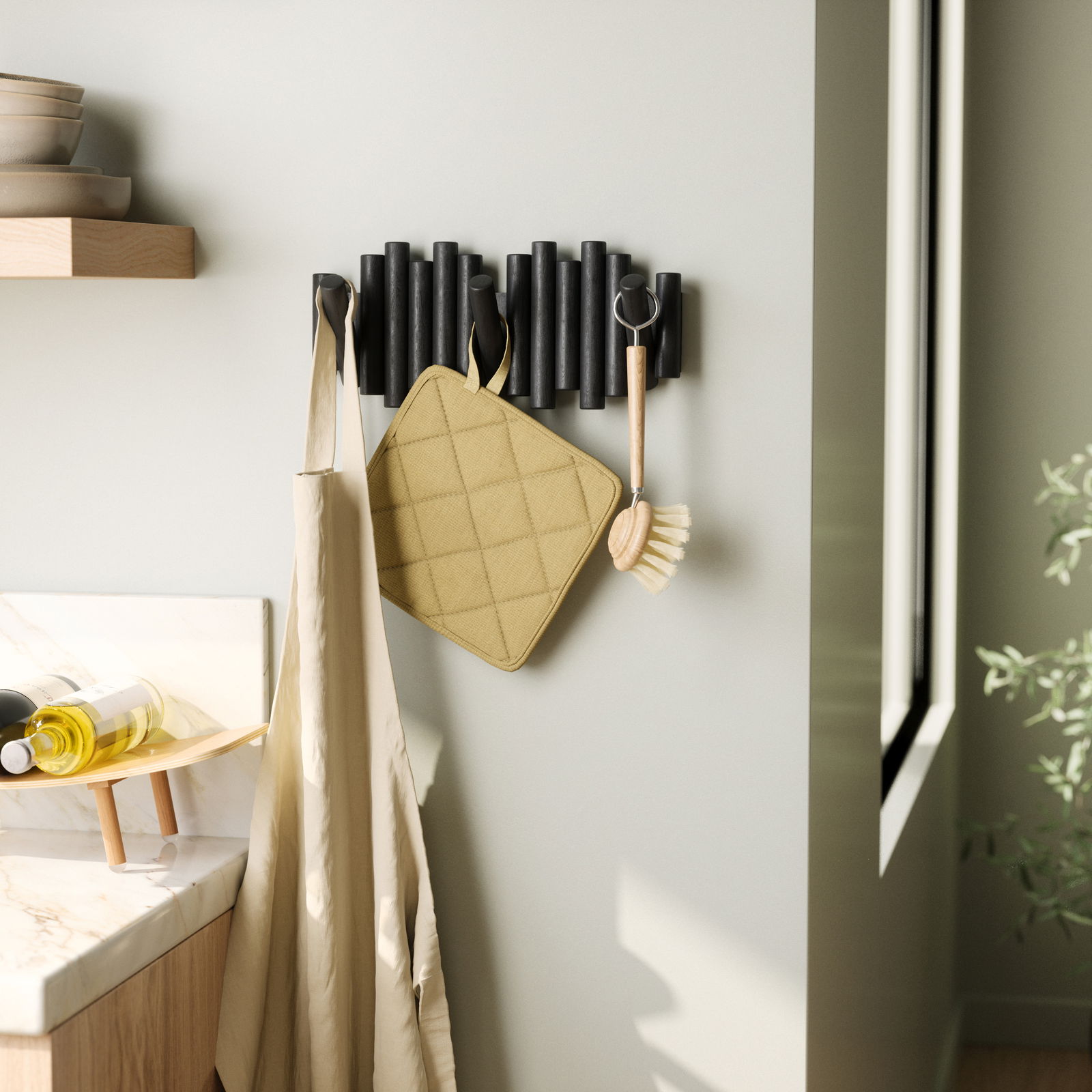 Moderne Wandgarderobe mit schwarzen Haken, an der ein beiger Stoffbeutel, ein gelber Topflappen und eine Spülbürste hängen. Perspektive von der Seite.