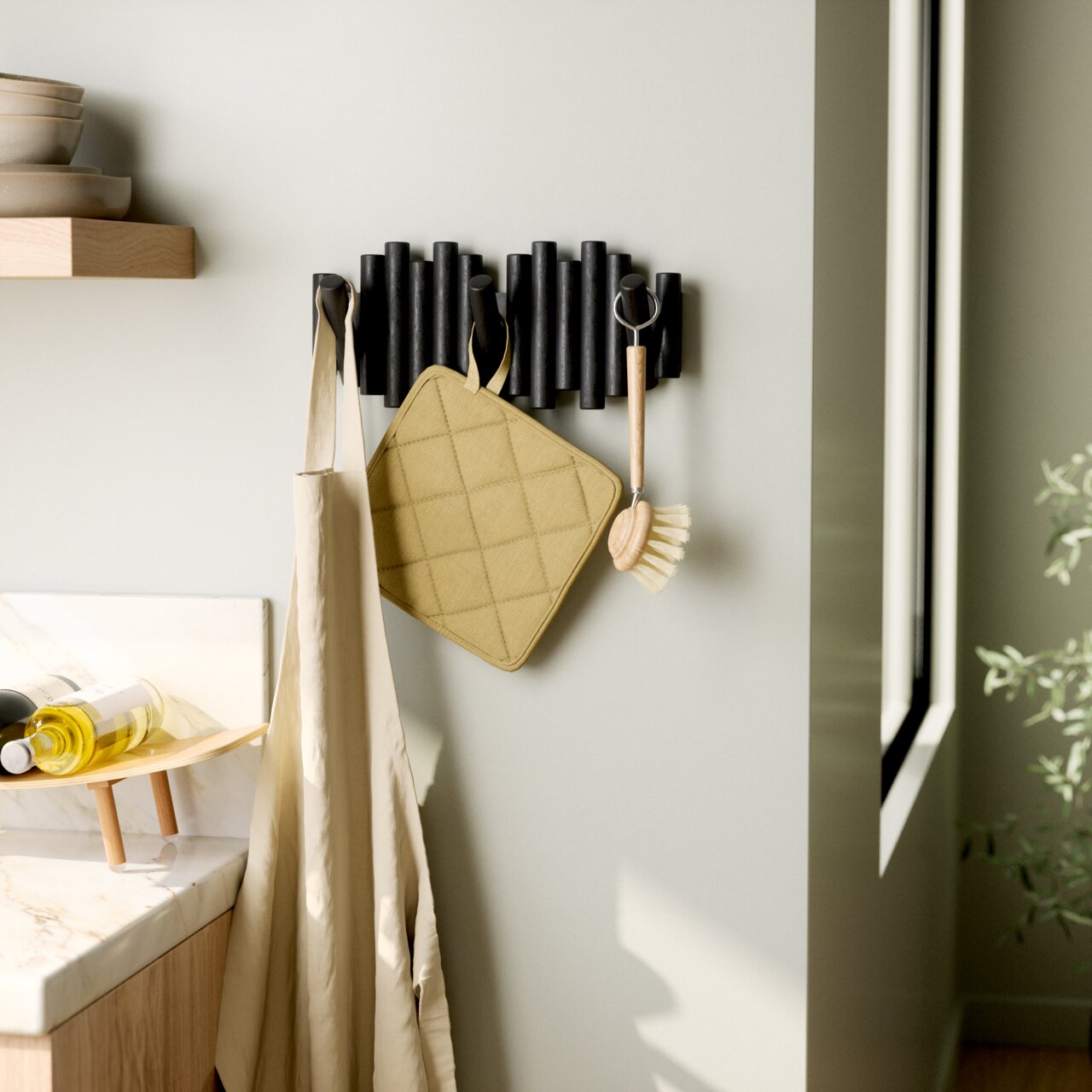 Moderne Wandgarderobe mit schwarzen Haken, an der ein beiger Stoffbeutel, ein gelber Topflappen und eine Spülbürste hängen. Perspektive von der Seite.