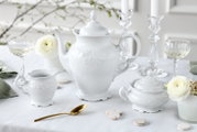 Weiße Porzellan-Teekanne mit Deckel im Barockstil, frontal aus leicht seitlicher Perspektive auf gedecktem Tisch, flankiert von Milchkännchen und Zuckerdose sowie Kristallgläsern und weißen Blumen.