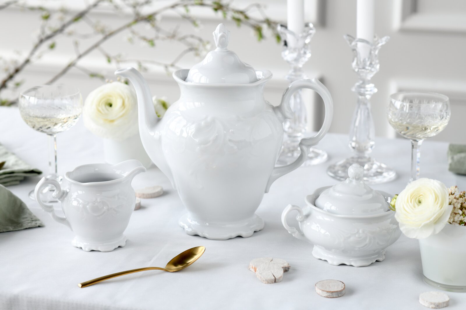 Weiße Porzellan-Teekanne mit Deckel im Barockstil, frontal aus leicht seitlicher Perspektive auf gedecktem Tisch, flankiert von Milchkännchen und Zuckerdose sowie Kristallgläsern und weißen Blumen.